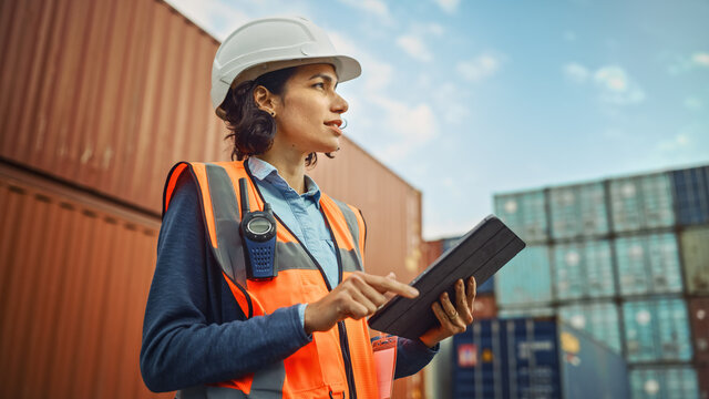 Smiling Portrait of a Beautiful Latin Female Industrial Engineer in White Hard Hat, High-Visibility Vest Working on Tablet Computer. Inspector or Safety Supervisor in Container Terminal.