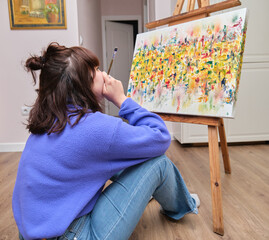 Young woman thinking and painting on canvas with oil paints at home. Artist drawing on easel sitting on the floor.