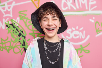 Happy teenage biy with braces on teeth wears stylish clothes with chains around neck has fun with friends poses against colorful graffiti wall as example of modern art. Youth and lifestyle concept