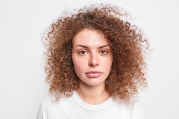 Portrait of serious millennial girl has healthy skin brown curly bushy hair dressed casually looks self confident at camera with calm face expression shows her natural beauty isolated on white wall
