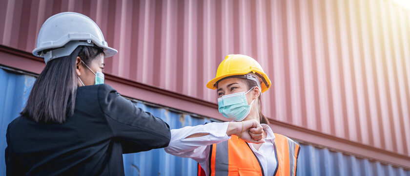 Famale manager and Engineer wearing face masks and greeting with elbow bump with shipping containers in the background. Coronavirus pandemic. Social distancing.