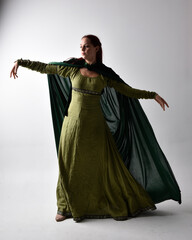 full length portrait of red haired girl wearing celtic, green medieval gown  and velvet cloak with shadowy backlighting. Standing pose isolated against a studio background.