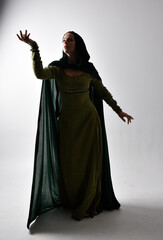 full length portrait of red haired girl wearing celtic, green medieval gown  and velvet cloak with shadowy backlighting. Standing pose isolated against a studio background.