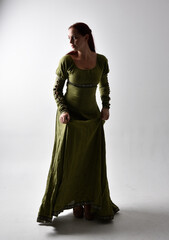 full length portrait of red haired girl wearing celtic, green medieval gown  with shadowy backlighting. Standing pose isolated against a studio background.