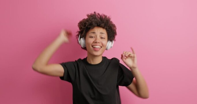 Joyful curly haired woman jumps and dances catches every bit of music wears casual black t shirt enjoys song in wireless headphones isolated over pink background. Amused entertained teenage girl