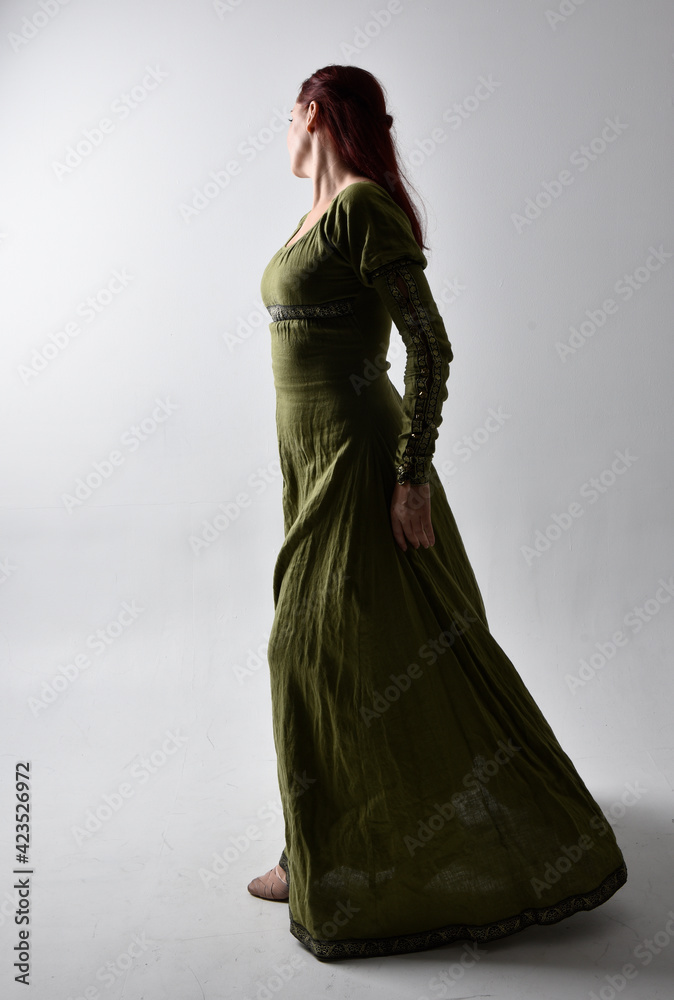 Poster full length portrait of red haired girl wearing celtic, green medieval gown  with shadowy backlighting. Standing pose isolated against a studio background.