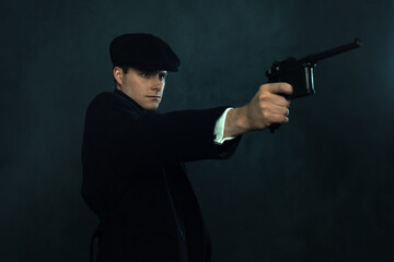 Threatening vintage man in a dark striped suit and a cap in front of a dark wall pointing a handgun.
