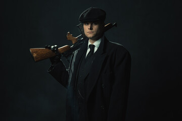 Threatening vintage man in a dark suit and cap in front of a dark wall holds a shotgun over his...