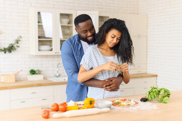 Loving black husband hugs wife who preparing his favorite food - homemade delicious pizza, hungry man waiting for a tasty yummy lunch, watches his wife decorates the dough with greenery and filling