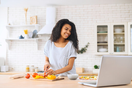 Portrait of nice attractive lovely biracial girl cooking vegs using laptop, watching tutorial video master-class, using knife to cut vegetables on board, making a pizza, standing on the modern kitchen - Powered by Adobe