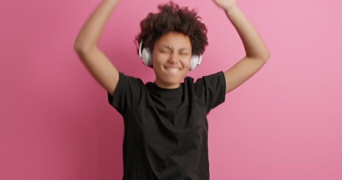 Beautiful joyful millennial girl with curly hair keeps arms raised feels happy wears casual black t shirt listens music via wireless headphones expresses positive emotions isolated on pink background