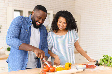 Young happy family prepares their first dish together in a new apartment, husband cuts cheese for pizza, stand at the table top with with many vegetables and ready-made dough smeared with tomato paste