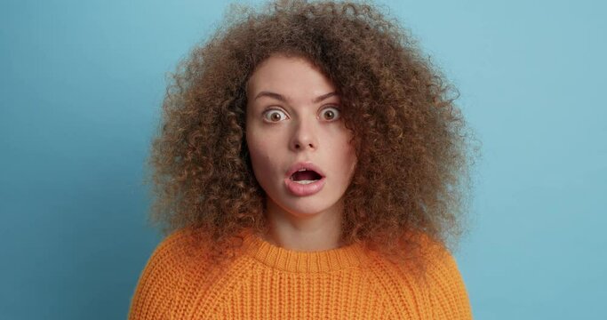 Startled curly young European woman gazes very surprisingly at camera being amazed by something says its unbelievable dressed in orange sweater isolated over blue background. Wow how incedible