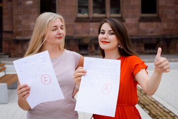 Two female friends with different test results sitting outdoors in the campus. Blond and brunette girls express their emotions after seeing their marks of the exam.