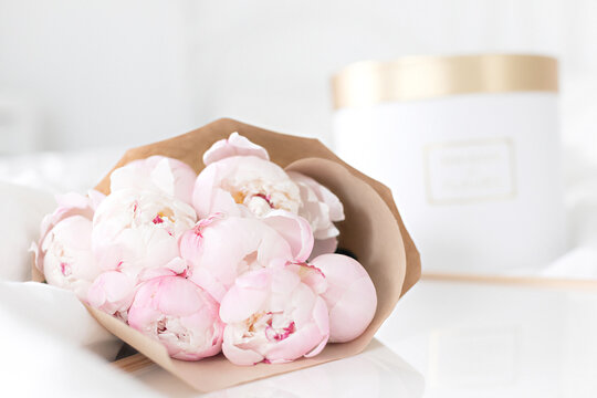 Gold gift box and white frame mockup on the bed. Bouquet of pink peonies in craft packaging. Scandivanavian white interior.