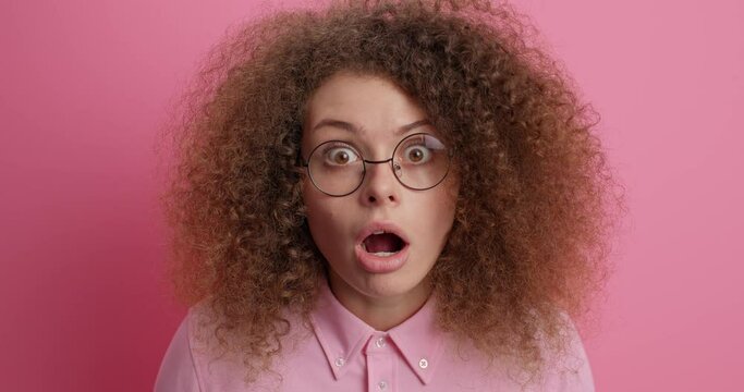 Beautiful curly haired young woman gasps amazed says wow and stands fascinated checks out promo offer stares through transparent glasses poses against pink background. Awesome advertisement.