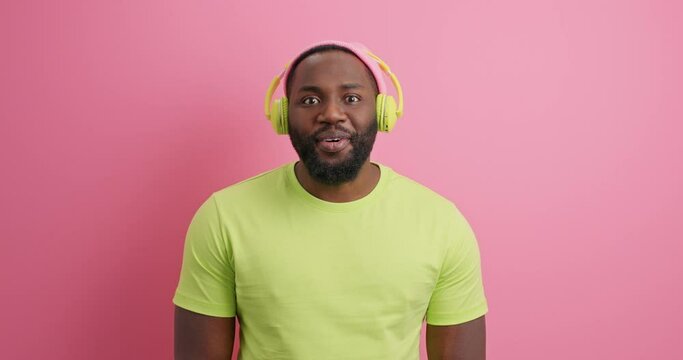 Surprised Afro American guy says wow cannot believe his eyes wears casual t shirt listens favorite music via wireless headphones isolated over pink background. People lifestlye hobby concept
