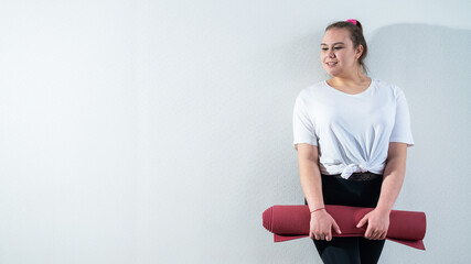 Young fat caucasian woman holding a sport mat. Charming plus size model in sportswear stands on a...