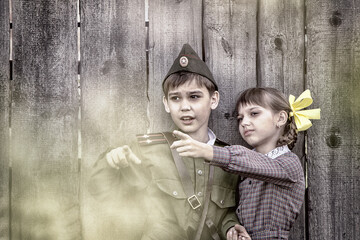 Postcard, stylized as vintage for the Victory Day. A boy in a military uniform and a girl in an old dress are standing near an old fence. The theme of May 9, Victory Day in Russia.
