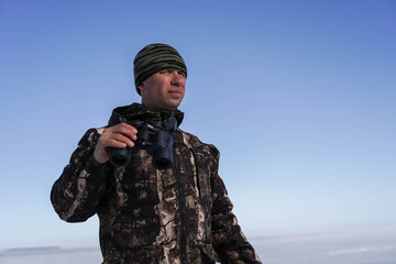 a man in military clothes holds binoculars in his hand and looks.  In the background snowy steppe and blue sky.