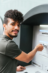 Obraz premium latin american young man looking at camera and smiling while using an ATM