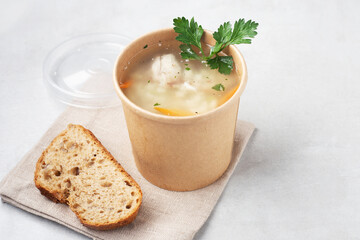chicken soup in a paper cup on a gray table with herbs, a piece of bread