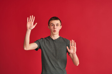 surprised, frightened young man stands in a mime pose with his arms outstretched on a red background, as if looking through a glass. The concept of a hopeless situation, problems, fear, claustrophobia