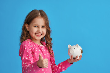 Fototapeta na wymiar Happy savings. A little cute girl with a piggy bank on a blue background. The child smiles happily and hugs him tightly and shows him a thumbs up. The concept of saving money for a dream.