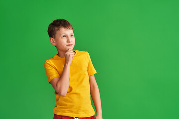pensive 8-year-old boy in a yellow T-shirt stands and thinks on a green background. The child ponders his decision, reflects on the correct answer. Copy the space.