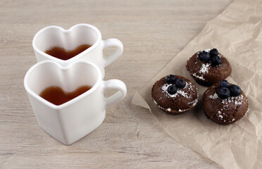 Two mugs in the shape of a heart with tea and muffins on baking paper on a wooden background. Tea party for two