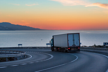 Truck with refrigerated semi-trailer on a mountain road with the sea and sunrise on the horizon.