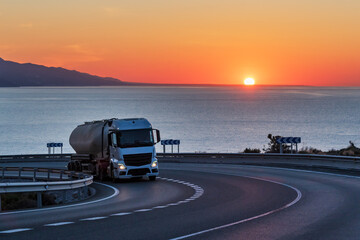 Truck with semi-trailer for the transport of bulk cement on a mountain road with the sea and the sun in the background on the horizon.