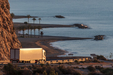 Truck with refrigerated semi-trailer on a mountain road that goes down to a beach with palm trees.
