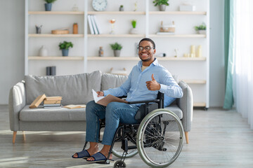 Cheerful handicapped black guy in wheelchair holding notebook, showing thumb up gesture at home