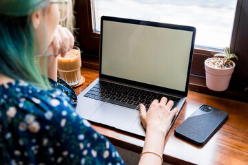 Close-up of female hands using laptop and smartphone at coffee shop with white mockup screen, women's hands typing on laptop keyboard, side view of a woman using computer in cafe