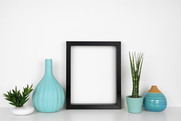 Mock up black frame with houseplants and blue decor. White shelf against a white wall. Copy space.