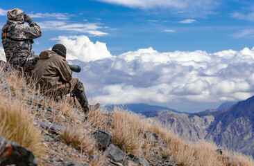 Two men in camouflage on the slope are watching in the optics in the mountains.