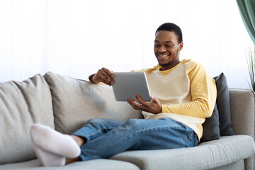Positive black man reclining on sofa, using digital tablet