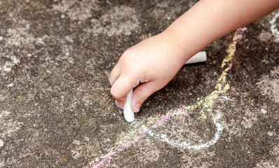 Little child's hand, girl holding white chalk, drawing, writing on the concrete outside. School age...
