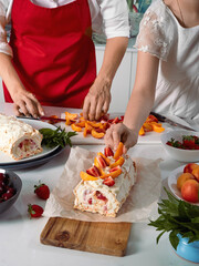 Meringue roll with a delicate air cream, mint and fruit filling of strawberries and peaches. Pavlova summer sweet dessert. Summer baking. Copy space. Mom and daughter prepare meringue roll.