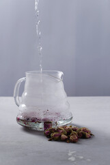 The concept of brewing tea from buds and rose petals. Hot water is poured into the glass kettle to make tea. The kettle stands on a gray concrete vertical background, selective focus