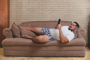 A typical couch potato - A man in his underwear laying on the sofa and watching videos, playing...