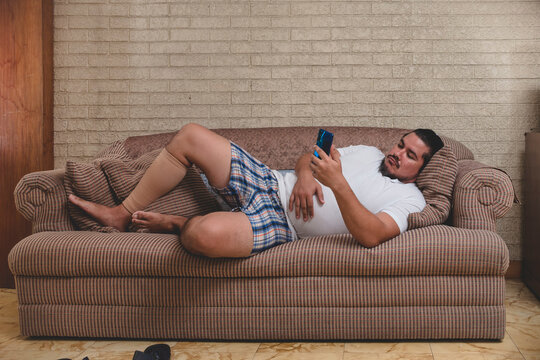 An unemployed and lazy man watching videos, playing mobile games or on social media on his cellphone all day while lying on the couch recovering from a calf injury.