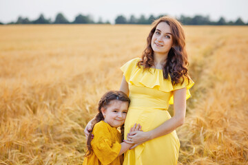 Naklejka na ściany i meble Pregnant mother and daughter in a wheat field at sunset. Gentle family hugs of mother and child in nature.