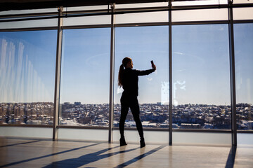 Fototapeta na wymiar A beautiful girl takes a selfie on her phone against the background of panoramic windows in a skyscraper