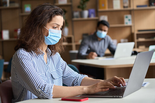 Latin girl student wearing face mask studying on laptop. Hispanic professional woman employee in facemask working sitting at table in modern office coworking space keeping safe social distance.