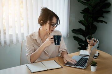 Portrait of beautiful accountant sitting at desk with interior drinking hot beverage holding cup with coffee looking at laptop while video conference.