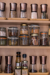 Glass piled jars and manual copper elegant mills full of spices, salt, aromatic plants over wooden kitchen selves.