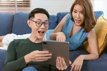 Excited Asian couple man and woman using tablet together on sofa at home. family lifestyle concept.