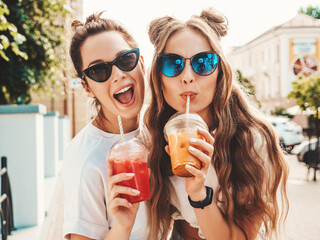 Two young beautiful smiling hipster female in trendy summer clothes. Carefree women posing outdoors.Positive models holding and drinking fresh cocktail smoothie drink in plastic cup with straw